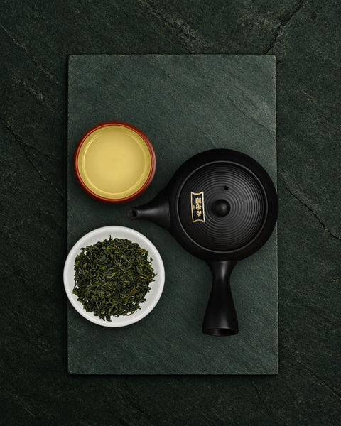A black ceramic teapot with gold calligraphy on the lid, displayed on a dark slate tray alongside a white bowl of kamairicha loose green tea leaves and a small red-rimmed cup of yellow tea, shot from a top-down angle.