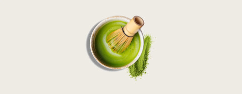A top-down view of a small bowl filled with bright green matcha tea, with a bamboo whisk resting inside the bowl and a small pile of green matcha powder beside it on a light warm gray background.