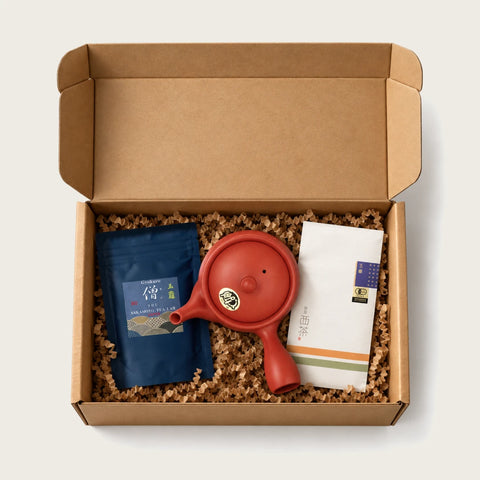 An open kraft cardboard gift box filled with brown packing paper and tea accessories, including a red teapot, a blue tea pouch, and a white tea packet.